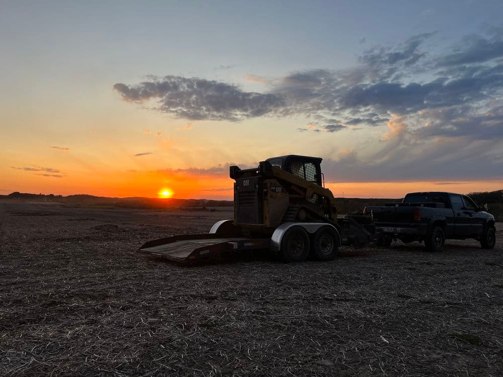 Skid steer loader on trailer with truck at sunset over the horizon in a farm field.