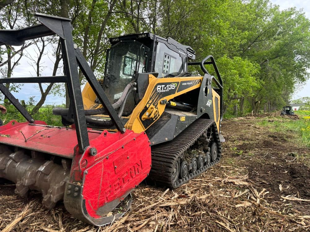 Bobcat RT 133 compact track loader with forestry attachment clearing land in wooded area.