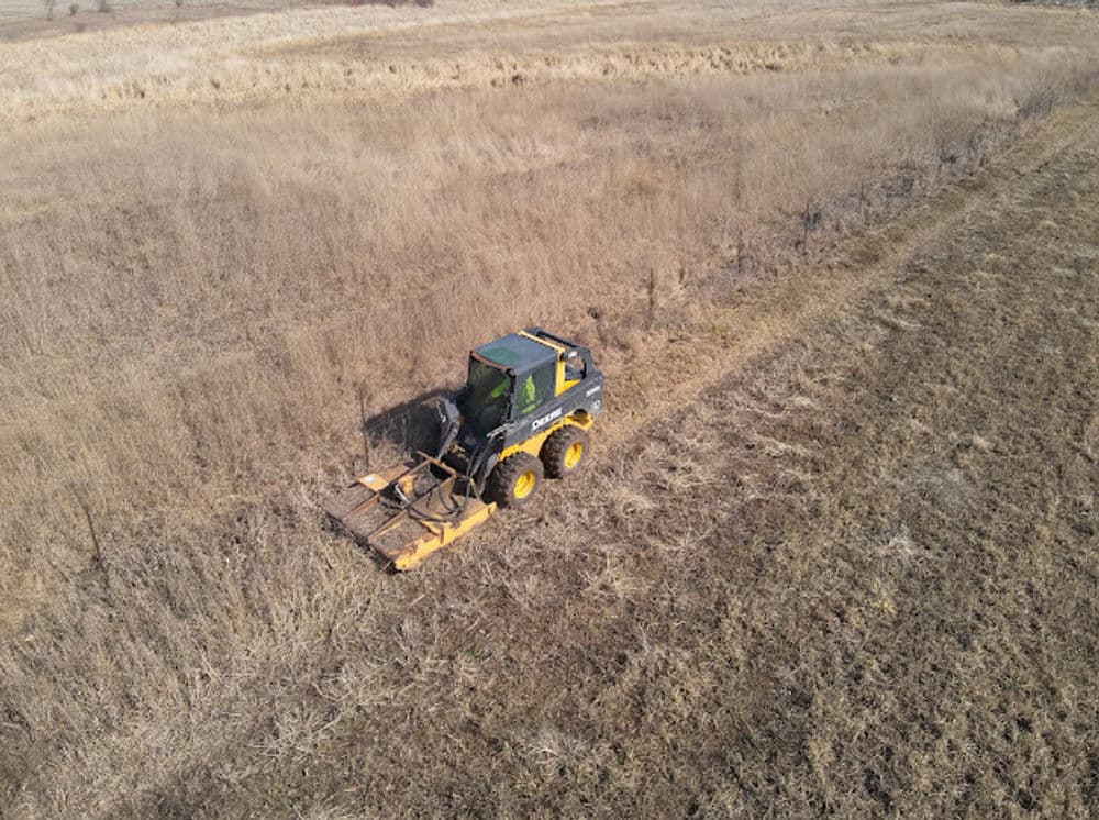 A tractor clearing overgrown grass in a dry, open field during daylight.