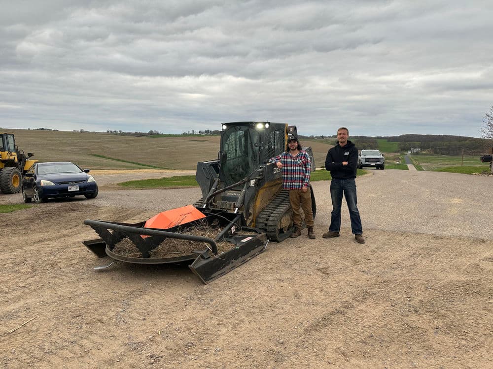 Two men stand beside a large skid steer loader on a gravel road with fields in the background.
