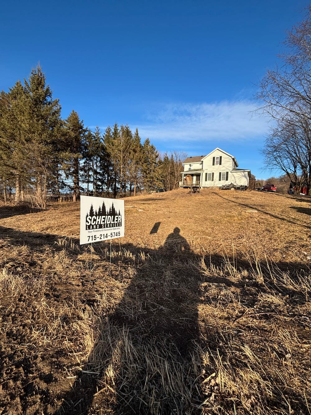 For sale sign in front of a house on a grassy lot surrounded by trees under a clear blue sky.