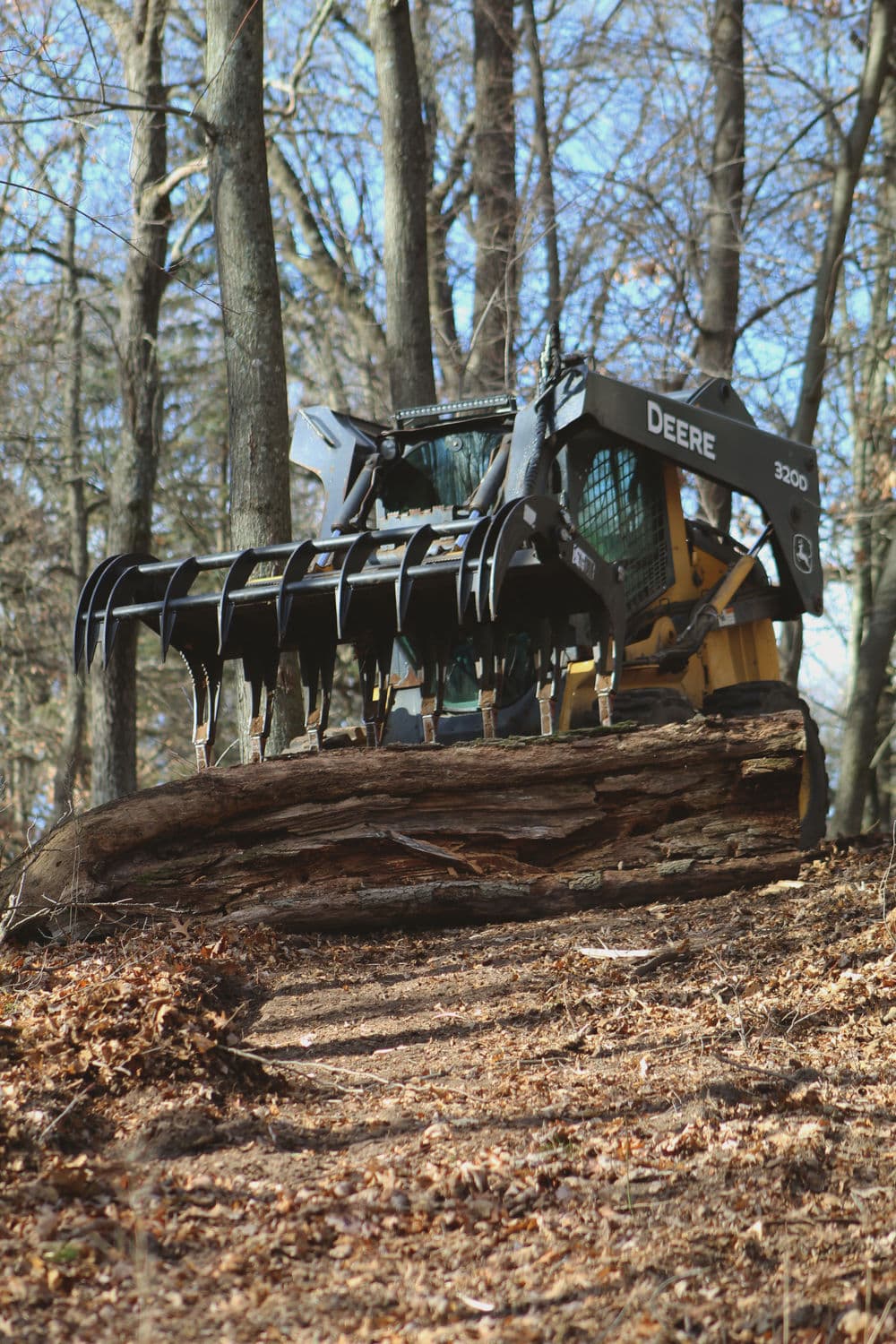 John Deere 320G skid steer loader lifting a log on a forested trail in winter.