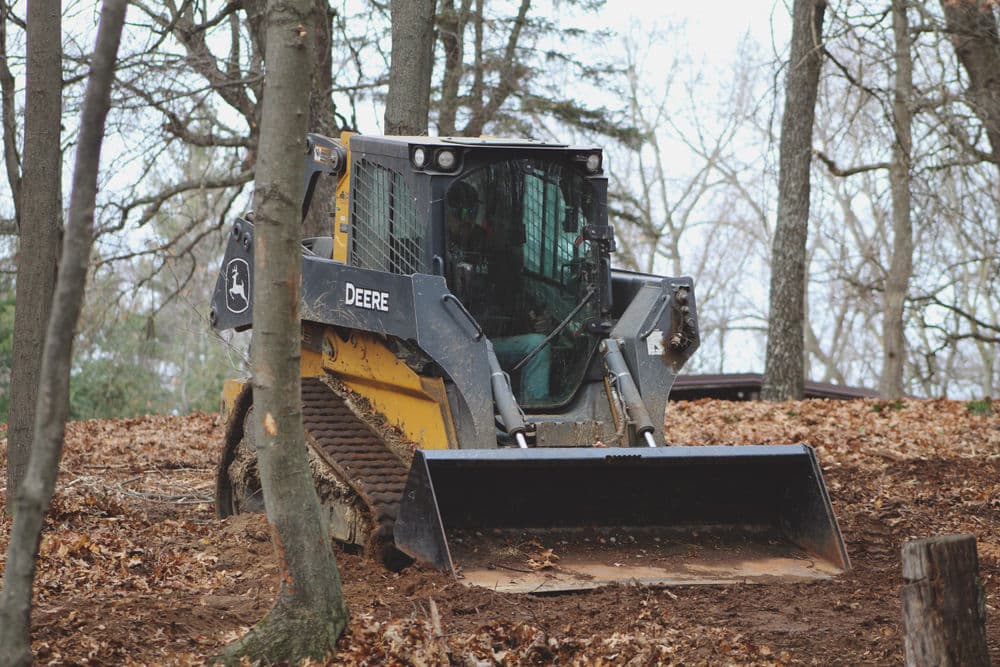 John Deere skid steer loader operating in a wooded area clearing leaves and debris.