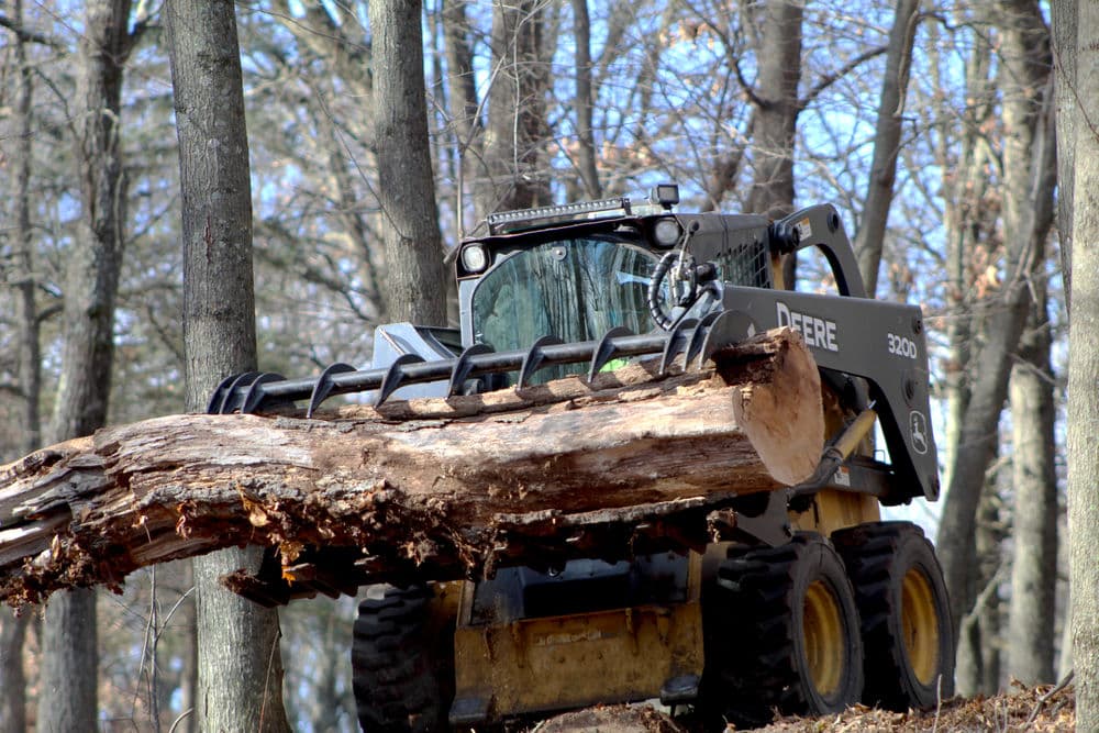 John Deere skid steer loader lifting a large log in a wooded area.